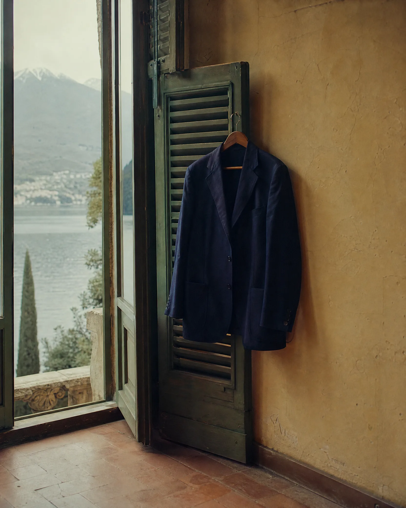 A navy blazer on a wooden hanger, hung from a green shutter beside a window over the lake.