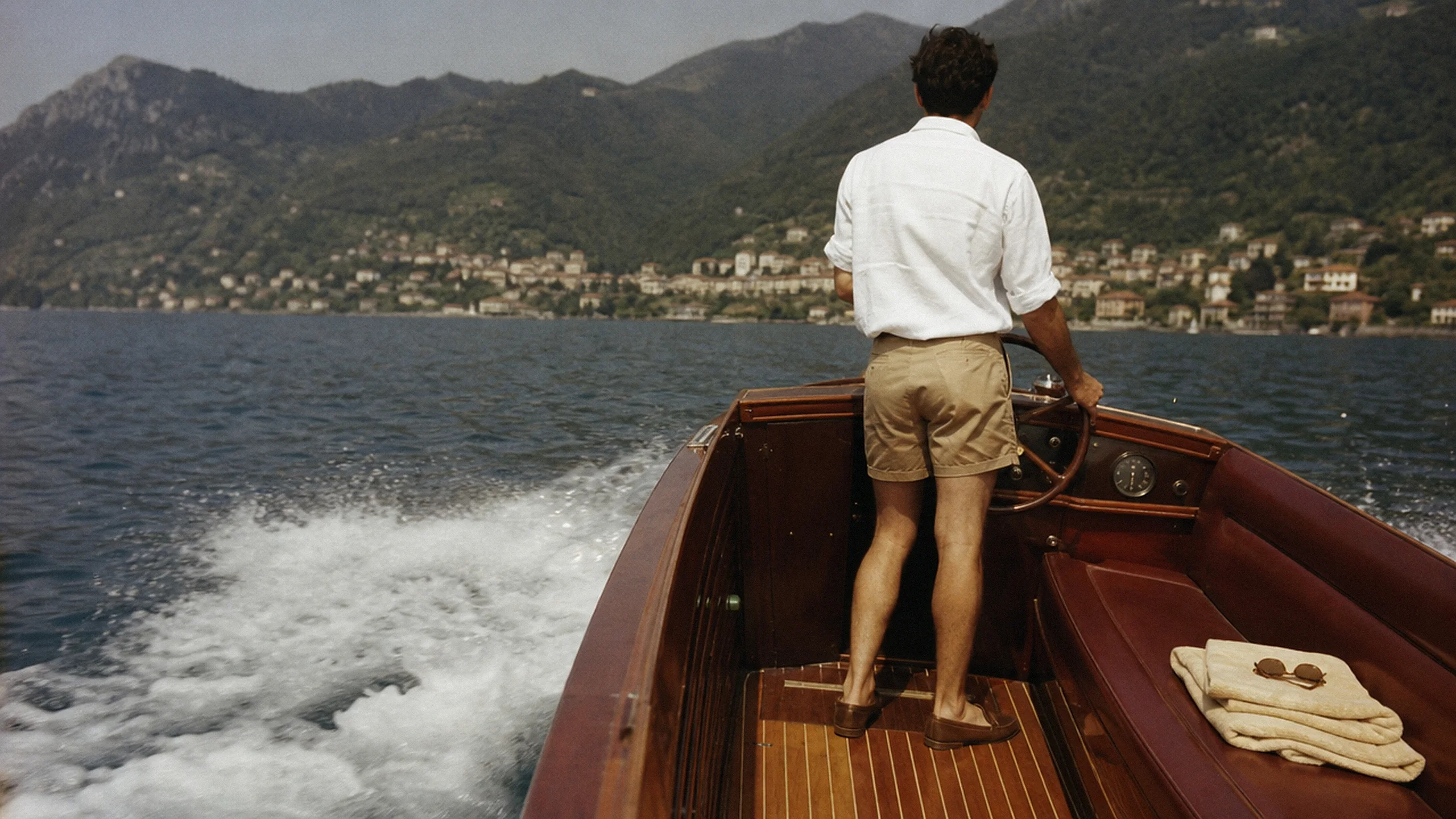 A man at the wheel of a varnished wooden motorboat, seen from behind, in a white linen shirt and tan shorts, crossing the lake toward a village.