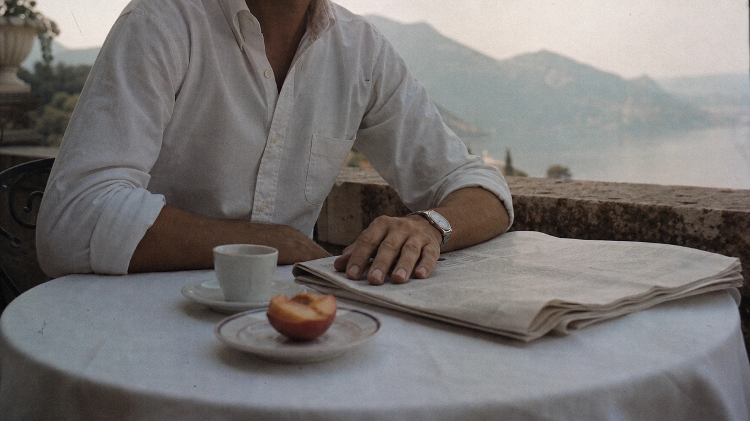 A man at a small table on a terrace above the lake, shown from the chest down: a white linen shirt with the sleeves rolled, an espresso, a folded newspaper and half a peach.