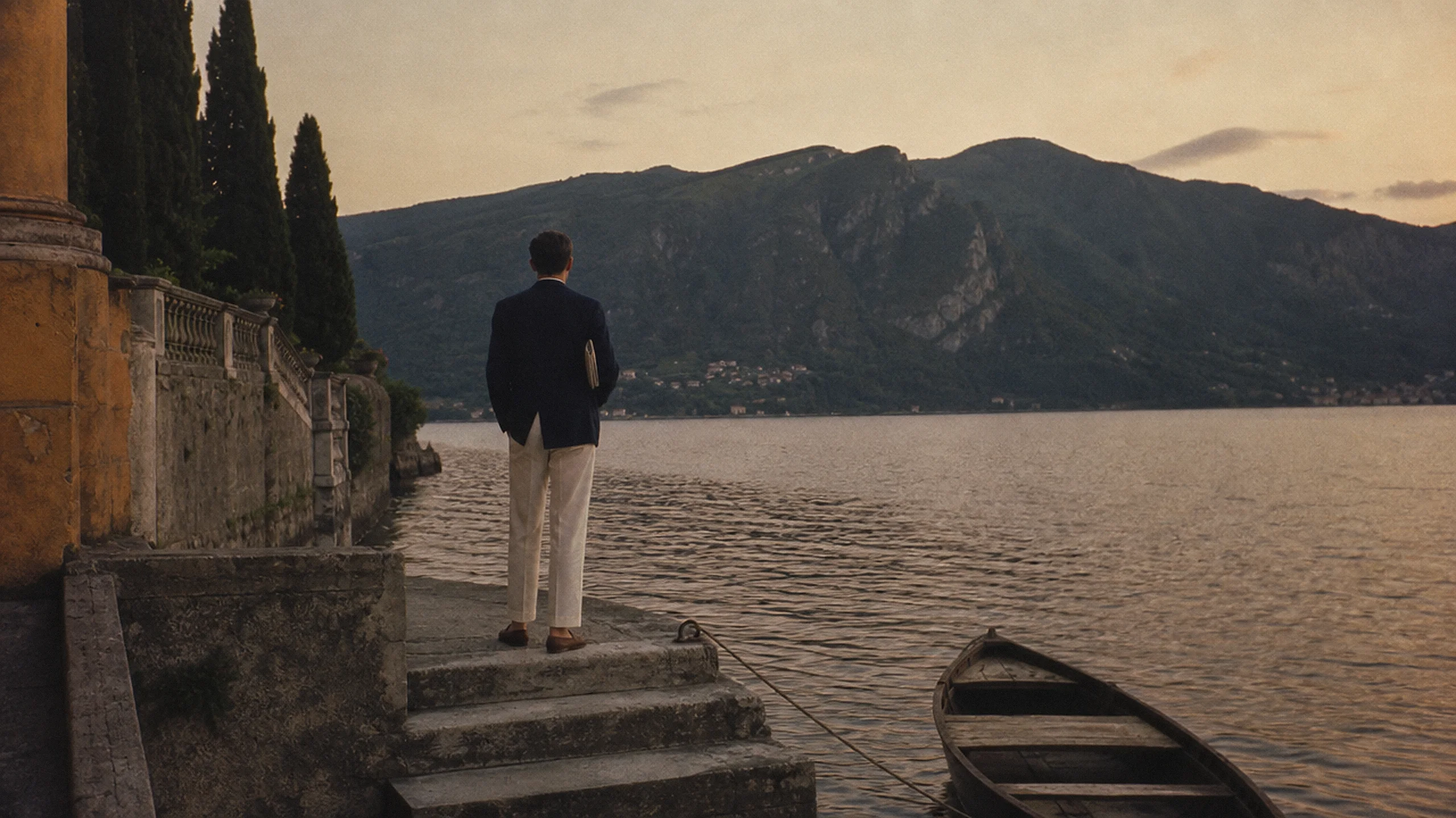 A man in a navy blazer and cream trousers, seen from behind, on the boat steps of a villa at dusk, looking across the lake.