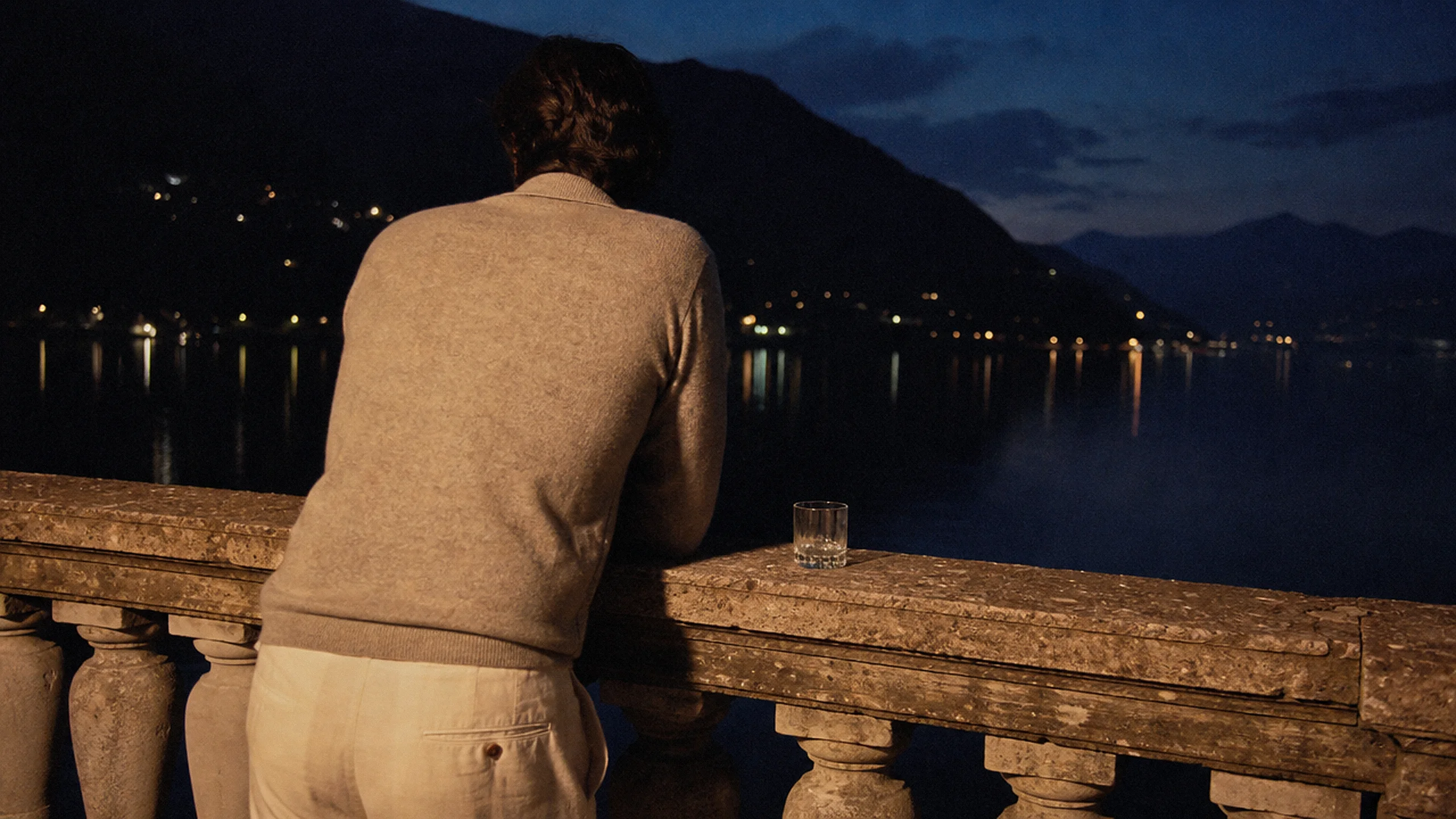 A man leaning on a stone balustrade at night, seen from behind, in an oatmeal quarter-zip sweater and cream trousers, with the lights of a village across the water.