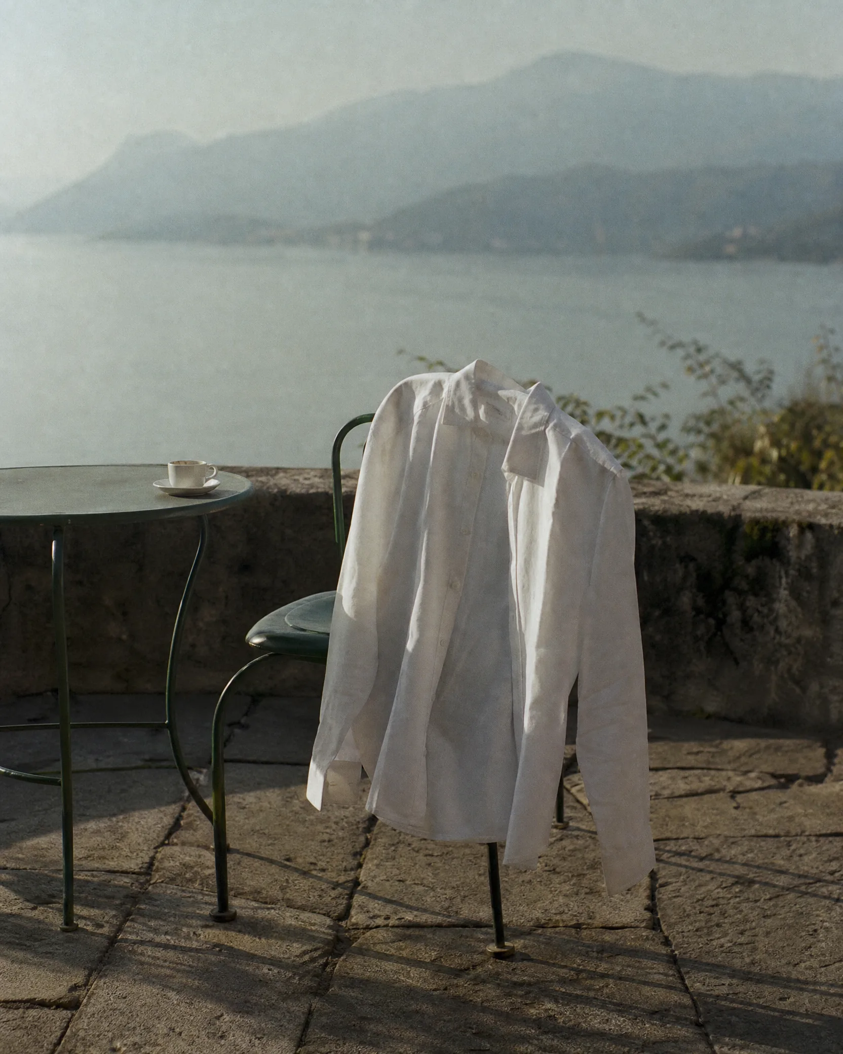 A white linen shirt hung over the back of a green iron chair on a terrace above the lake, an espresso on the table beside it.